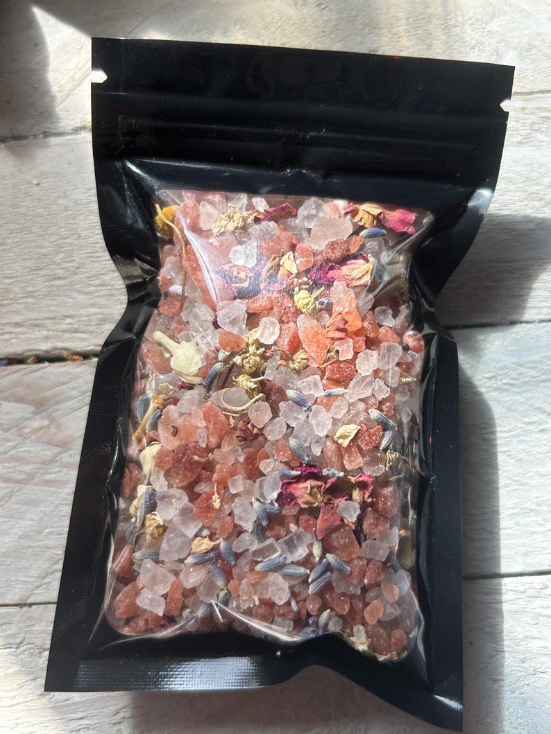 May include: A black, resealable bag filled with a mixture of bath salts. The salts are a mix of pink, white, and clear crystals, with dried lavender and rose petals. The bag is sitting on a white wooden surface.