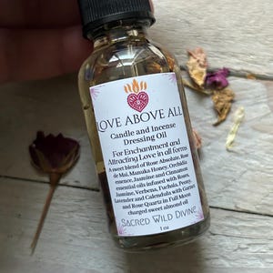 May include: A clear glass bottle of "LOVE ABOVE ALL" candle and incense dressing oil. The label features a heart and flame design. The bottle contains a golden liquid and is topped with a black dropper. The text on the label describes the ingredients.