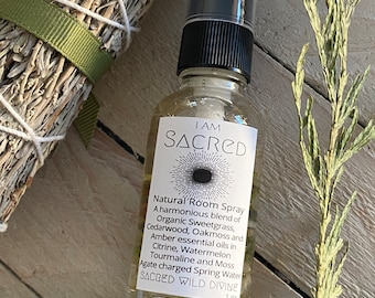 May include: A clear glass bottle with a black spray top and a white label that reads "I AM SACRED" and lists the ingredients of the natural room spray. The bottle is sitting on a wooden surface with a bundle of dried sage and a bundle of dried grass.