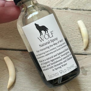 May include: A clear glass spray bottle with a black cap and a white label featuring a wolf silhouette and the text "WOLF Natural Spray." The bottle contains a dark liquid. Two off-white, curved objects are next to the bottle. The label also includes the text "SACRED WILD DIVINE."