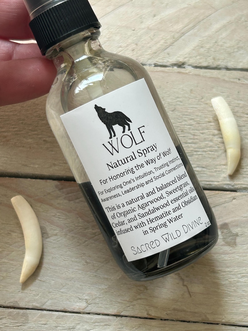 May include: A clear glass spray bottle with a black cap and a white label featuring a wolf silhouette and the text "WOLF Natural Spray." The bottle contains a dark liquid. Two off-white, curved objects are next to the bottle. The label also includes the text "SACRED WILD DIVINE."