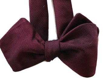 May include: A burgundy bow tie made from a textured fabric. The bow tie is tied and ready to wear.