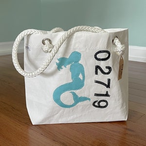 May include: White canvas tote bag with a rope handle and a blue mermaid silhouette. The bag has the number "91720" printed in black.