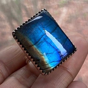 May include: A silver ring with a large, rectangular, iridescent blue labradorite gemstone. The stone has a dark blue, green, and gold color.