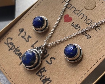 Puede incluir: Un conjunto de collar y aretes de plata con piedras de lapislázuli azules. El collar tiene un colgante con una piedra redonda y los aretes tienen pequeñas piedras redondas. La etiqueta dice "Hecho a mano con amor" y "925 Lapis Piedra de nacimiento de septiembre".