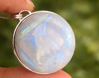 May include: A round, iridescent moonstone pendant with a silver-colored bezel. The moonstone displays a milky white base with flashes of blue and yellow. The pendant is suspended from a delicate silver chain, creating a simple and elegant necklace.