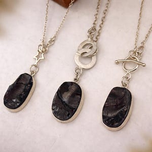 May include: Three silver necklaces with dark, oval pendants. Each pendant is set in a silver bezel. The necklaces have different chain styles and clasps, including a star, a double ring, and a toggle clasp.