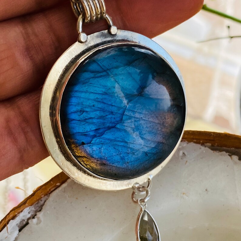 "Indulge in the celestial beauty of the Earth Karma Celestial Blue Flash Labradorite Necklace, resembling a round cosmic galaxy, set in sterling silver, and exuding timeless elegance, accentuated by a moonstone dangle."