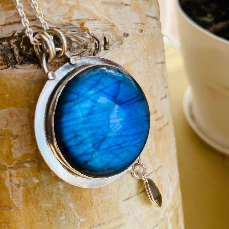 "Admire the detail of the EarthKarma Hand Carved Raw Cabochon Labradorite Pendant, capturing the essence of a round blue moon in the night sky, set in sterling silver, for a bohemian touch, with an accompanying moonstone dangle."