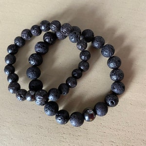 Puede incluir: Dos pulseras de cuentas de roca volcánica negra. Las pulseras están hechas de pequeñas cuentas redondas con una superficie rugosa y texturizada. Una pulsera tiene una sola cuenta negra, más grande y lisa.