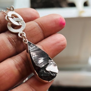 May include: A silver necklace with a teardrop-shaped pendant featuring a black, metallic, textured stone.