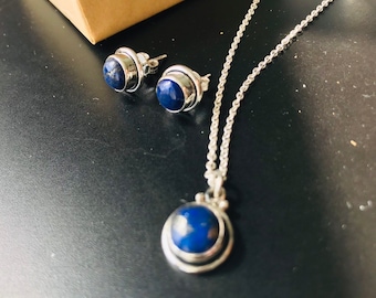 Puede incluir: Un conjunto de collar y pendientes de plata con piedras de lapislázuli azules. El collar tiene una cadena delicada y un colgante con una piedra redonda.