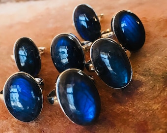 May include: Seven silver rings with oval-shaped blue labradorite gemstones. The rings are arranged in a semi-circle on a wooden surface.