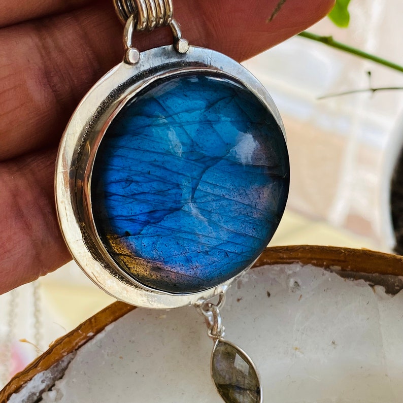 "Step into the extraordinary with the Earth Karma Beyond Blue Labradorite Pendant, featuring a round design reminiscent of a cosmic galaxy, elegantly set in sterling silver, offering timeless elegance, paired with a moonstone dangle."
