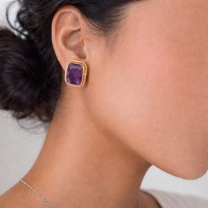 May include: A close-up of a gold-colored square earring with a large, rectangular, deep purple gemstone. The earring has a textured, rope-like border. The earring is worn on a person's ear.