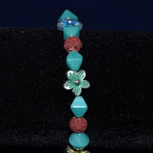 Puede incluir: Un brazalete de cuentas turquesa y rojo con un dije de flor de plata. El brazalete está sobre un fondo negro.