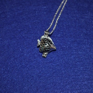Puede incluir: Un collar de plata con un dije en forma de pez y una jaula. La jaula está abierta y parece estar vacía.