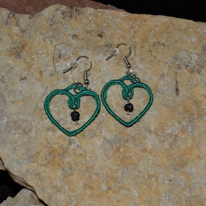 Puede incluir: Un par de pendientes de corazón verde con cuentas negras que cuelgan de la parte inferior. Los pendientes están hechos de alambre y tienen un diseño delicado e intrincado.