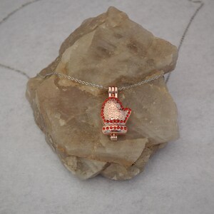 Könnte beinhalten: Eine Halskette mit einem roségoldenen Anhänger in Form einer roten, mit Edelsteinen besetzten Mütze. Der Anhänger hängt an einer silbernen Kette und liegt auf einem Stein.