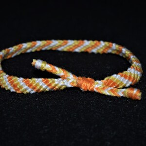 May include: A white, orange, and yellow friendship bracelet with a diagonal pattern.