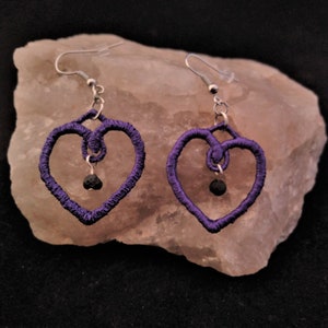 May include: Purple heart-shaped earrings with black lava rock beads. The earrings are made with wire and have a delicate, intricate design.