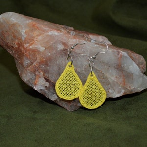 May include: A pair of yellow, teardrop-shaped earrings with an open-weave pattern. The earrings are suspended from silver-coloured ear wires. The earrings are shown against a natural stone background.