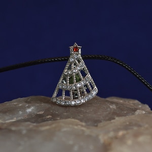 May include: A silver Christmas tree pendant with clear rhinestones and a red gemstone at the top. The pendant is hanging from a black cord.