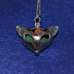 Puede incluir: Un collar difusor de aromaterapia de plata en forma de zorro con una cadena. El difusor tiene tres aberturas llenas de piedras verdes y marrones.