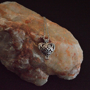 May include: A silver heart-shaped locket necklace with a delicate filigree design hangs against a rough, light brown rock.