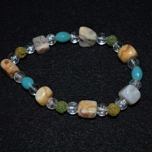 Puede incluir: Un brazalete hecho con cuentas de ágata marrón, blanco y azul, cuentas de vidrio transparentes y pequeñas cuentas de lava verde.