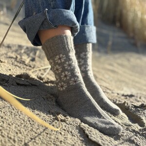 Puede incluir: Un par de calcetines grises de punto con un estampado floral en blanco y rojo. Los calcetines se llevan con vaqueros azules, con los bajos doblados en los tobillos. La imagen está tomada al aire libre sobre una superficie arenosa.