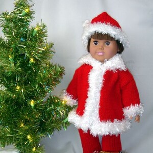 May include: A doll dressed in a red and white Santa suit with a white fur trim. The doll is holding a brown sack filled with wrapped gifts. The doll is standing in front of a green Christmas tree with lights.