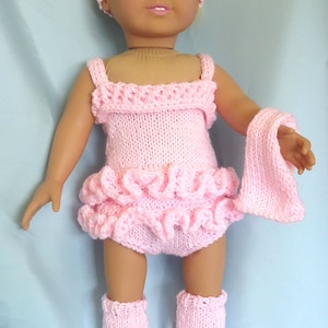 May include: A pink knitted doll outfit with a ruffled skirt, leg warmers, and headband. The doll is wearing white shoes.