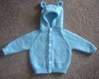 May include: A light blue knitted baby cardigan with a hood and bear ears. The cardigan has a button closure and is made of a soft, comfortable material.