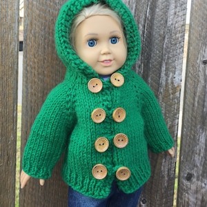 Könnte beinhalten: Grüne gestrickte Kapuzenjacke mit Holzknöpfen für eine Puppe. Die Jacke hat eine Kapuze und lange Ärmel.