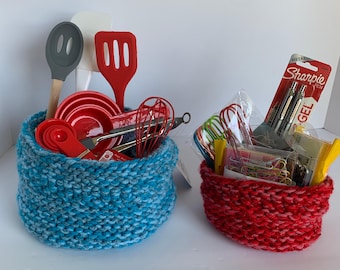May include: Two handmade, knitted baskets filled with kitchen and office supplies. The blue basket contains red kitchen utensils, while the red basket holds pens, paperclips, and other office items. The baskets are made of thick yarn.