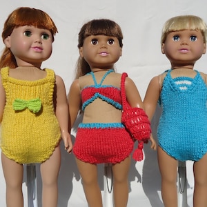 May include: Three dolls wearing different knitted swimsuits. The doll on the left is wearing a yellow swimsuit with a green bow. The doll in the middle is wearing a red and blue bikini with a matching red knitted bag. The doll on the right is wearing a blue swimsuit with white accents.
