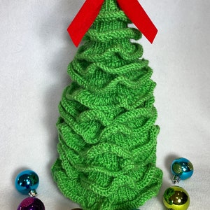 May include: A green knitted Christmas tree with a red velvet bow on top. The tree is surrounded by colourful baubles.