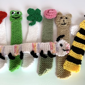 May include: A selection of handmade knitted bookmarks. Designs include a red heart, a green frog, a green shamrock, a pink rose, a tan bear, a striped caterpillar, and a yellow and black bee. Each bookmark is a unique, colourful design.