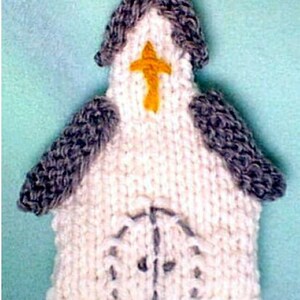 Könnte beinhalten: Ein weißer, gestrickter Kirchenanhänger mit einem grauen Dach und einem gelben Kreuz auf der Vorderseite. Die Kirche hat eine kleine Tür und Fenster.