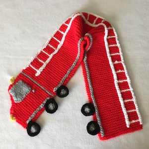 Op de afbeelding: Rode gebreide sjaal met een brandweerwagenontwerp. De brandweerwagen heeft een grijze ladder, zwarte wielen en gele details.