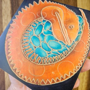 Può includere: Opera d'arte quadrata con un design di creatura stilizzata arancione e blu. L'opera ha uno sfondo nero e un bordo decorativo. La creatura è in posa circolare con un motivo dettagliato.