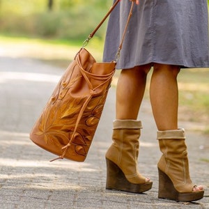 Puede incluir: Una mujer lleva un vestido camisa gris y botas de cuña marrones y sostiene un bolso de cuero marrón con un diseño floral.