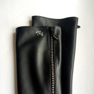 Könnte beinhalten: Schwarze Lederstiefel mit Reißverschlussdetail und einem dekorativen Schmuckstück an der Vorderseite. Das Schmuckstück ist ein silberfarbenes Metallstück mit einem kleinen, klaren Kristall.