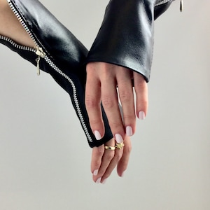 Könnte beinhalten: Ein Paar schwarze Lederfingerlose Handschuhe mit Reißverschlüssen auf der Rückseite der Hand. Die Handschuhe werden an beiden Händen getragen und die Finger sind sichtbar.