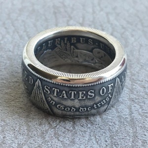 May include: A silver ring with a detailed design. The ring features the words "STATES OF AMERICA" and "In God we trust". The ring has a textured surface and a polished edge, showcasing a vintage aesthetic.
