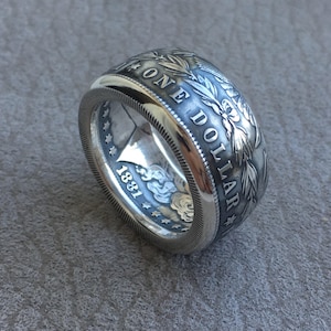 May include: A silver ring crafted from a coin, featuring the words "ONE DOLLAR" and the year "1881". The ring has intricate floral and leaf detailing around the edges, with a polished finish.
