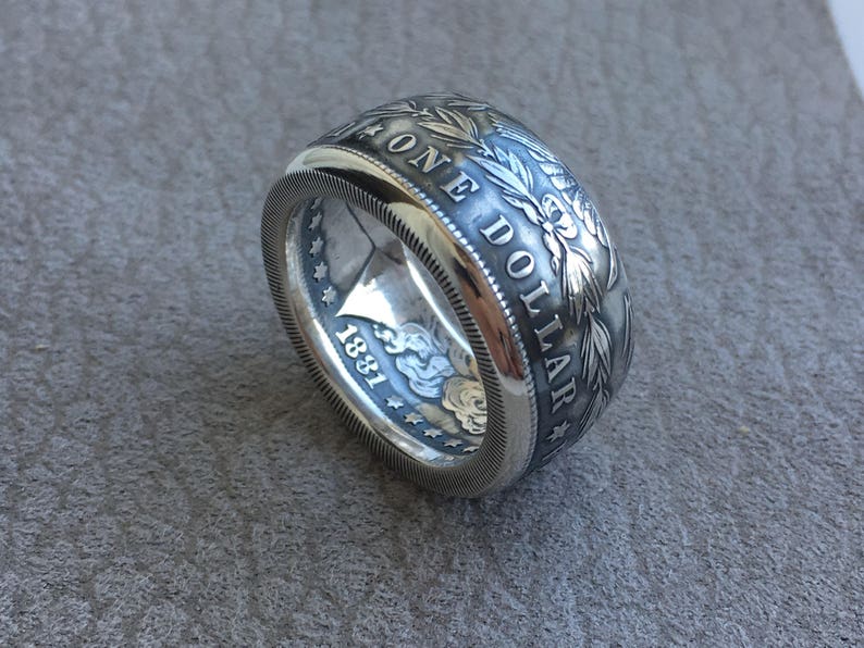 May include: A silver ring crafted from a coin, featuring the words "ONE DOLLAR" and the year "1881". The ring has intricate floral and leaf detailing around the edges, with a polished finish.