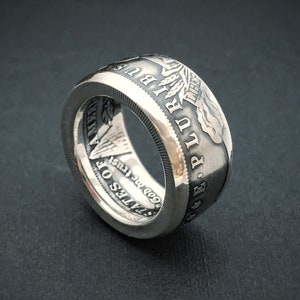 May include: A silver ring crafted from a coin, featuring engraved text and imagery. The ring displays the words "E PLURIBUS UNUM" and "STATES OF AMERICA" along with other details. The ring has a textured edge and a polished finish.