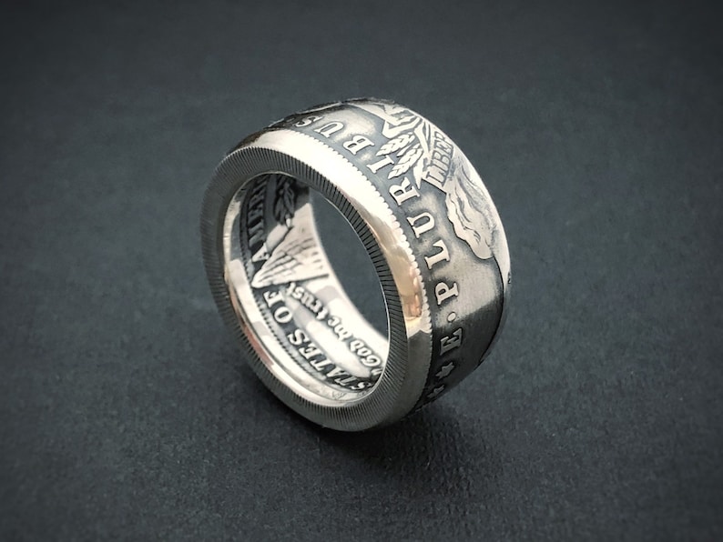May include: A silver ring crafted from a coin, featuring engraved text and imagery. The ring displays the words "E PLURIBUS UNUM" and "STATES OF AMERICA" along with other details. The ring has a textured edge and a polished finish.